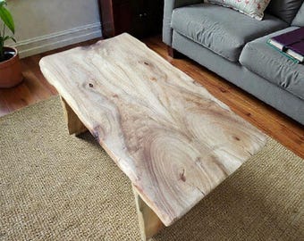 Puede incluir: Una mesa de centro rectangular de madera natural con borde vivo. La mesa mide aproximadamente 152 cm de largo y descansa sobre patas de madera sencillas. La veta de la madera es visible, mostrando tonos marrones claros y oscuros.