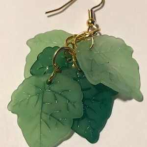 Puede incluir: Un par de pendientes colgantes de hojas verdes con ganchos dorados y anillos de conexión. Las hojas son translúcidas y tienen un delicado patrón de venas.