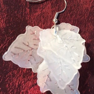 Może przedstawiać: Para białych, warstwowych kolczyków w kształcie liści z srebrnym haczykiem. Liście są półprzezroczyste i mają delikatny wzór żyłek.