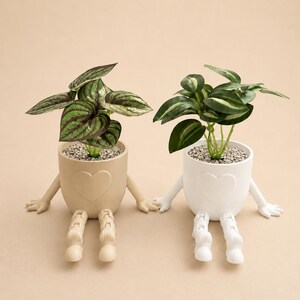Op de afbeelding: Twee keramische plantenpotten met armen en benen, één beige en één wit, elk met een hart in reliëf aan de voorzijde. De beige pot bevat een plant met donkergroene, gestreepte bladeren, terwijl de witte pot een plant met effen groene bladeren bevat.