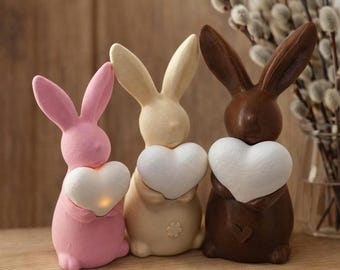 Può includere: Tre statuette di coniglietti pasquali rosa, crema e marroni, ognuno con un cuore bianco. Il cuore del coniglietto rosa si illumina. I coniglietti sono su una mensola di legno, con rami di salice sullo sfondo.