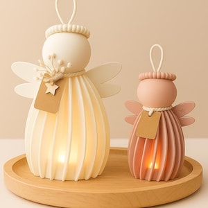 Op de afbeelding: Twee engelvormige decoratieve lampen, één crème en één roze, staan op een houten dienblad. Elk heeft een bolvormig hoofd, vleugels en een papieren label. De crèmekleurige engel is groter, met een stervormig label en gedroogde bloemen.