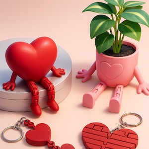Puede incluir: Una figura roja en forma de corazón con brazos y piernas se encuentra junto a una maceta rosa con un diseño de corazón y una planta pequeña. También se muestran dos llaveros rojos en forma de corazón, uno con una cadena de corazones y otro con un diseño texturizado.