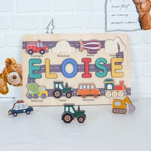 May include: Wooden puzzle featuring the name "Eloise" with colorful letters and transportation vehicles. Includes a fire engine, police car, airship, excavator, mixer truck, tractor, school bus, and train. A fun educational toy.
