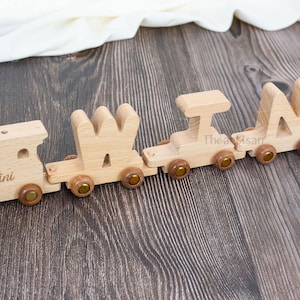 May include: A wooden toy train spelling out the name "MIA" in capital letters. The train cars are connected and feature wooden wheels with brass-colored accents. The train is made of light-colored wood and sits on a dark wood surface.