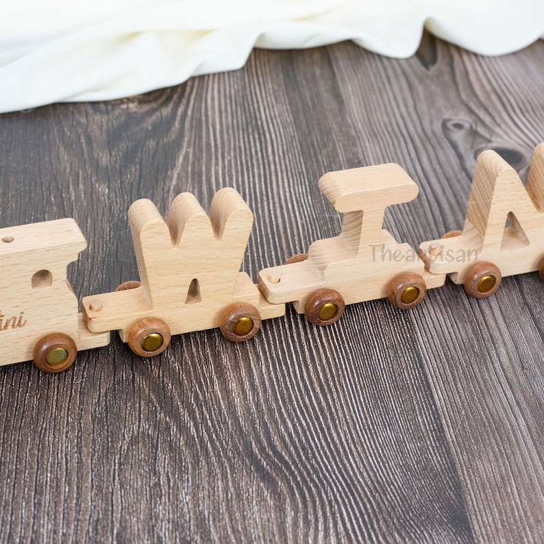 May include: A wooden toy train spelling out the name "MIA" in capital letters. The train cars are connected and feature wooden wheels with brass-colored accents. The train is made of light-colored wood and sits on a dark wood surface.