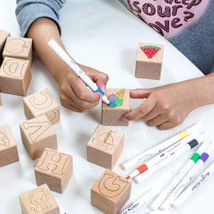 May include: Wooden alphabet blocks with engraved letters and images, including a lion, boat, and watermelon. A child is using acrylic markers to color the blocks, with several markers visible.