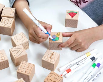 May include: Wooden alphabet blocks with engraved letters and images, including a lion, boat, and watermelon. A child is using acrylic markers to color the blocks, with several markers visible.