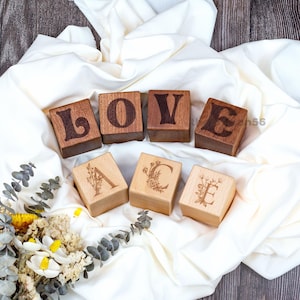 Peut inclure: Blocs de bois avec les lettres "LOVE" en marron foncé, et des blocs individuels avec des motifs floraux et les lettres "A", "C" et "E". Les blocs sont disposés sur un tissu blanc.
