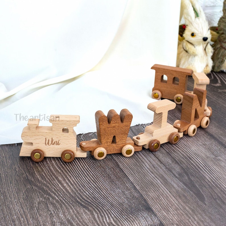 May include: Wooden toy train with four cars, each with a different design and color. The first car has the name "Wini" engraved on it. The train is made of light and dark brown wood with gold-colored wheels.