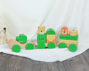 May include: A wooden toy train with green wheels and animal-themed blocks. The train features a crocodile, bear, and lion. The blocks are decorated with names and dates. The train is on a white cloth.