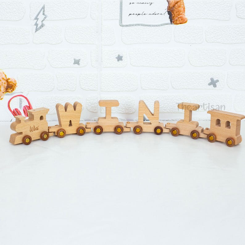May include: A wooden toy train spelling out the word "WIN". The train cars are connected and have brown wheels. The first car has the name "Utri" engraved on it. The background is a white brick wall.