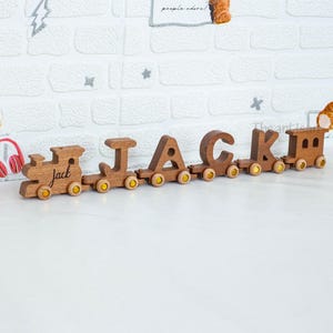 May include: Wooden train spelling out the name "JACK" in individual cars. The train is made of light brown wood with yellow wheels. The first car says "jack". The train is displayed on a white surface against a white brick wall.