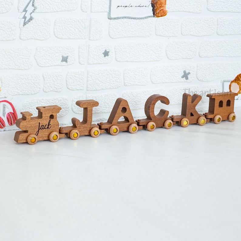 May include: Wooden train spelling out the name "JACK" in individual cars. The train is made of light brown wood with yellow wheels. The first car says "jack". The train is displayed on a white surface against a white brick wall.