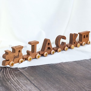 May include: A wooden toy train spelling out the name "Jack". The train cars are connected and feature the letters J, A, C, K, and a locomotive. Each car has small yellow wheels. The train is made of brown wood.