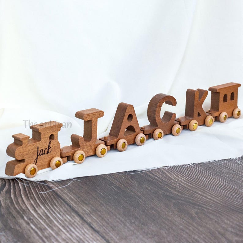 May include: A wooden toy train spelling out the name "Jack". The train cars are connected and feature the letters J, A, C, K, and a locomotive. Each car has small yellow wheels. The train is made of brown wood.