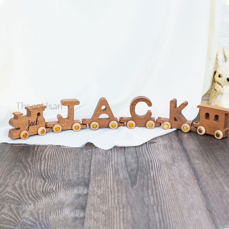 May include: Wooden train spelling out the name "JACK". The train cars are connected and have yellow wheels. The first car has the name "Jack" engraved on it. The train is made of a light brown wood and sits on a wooden surface.