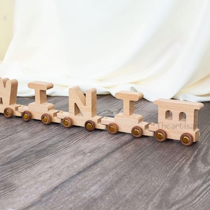 May include: A wooden toy train spelling out the name "MINI". The train is made of light-colored wood with gold-colored wheels. The train is on a wooden surface.
