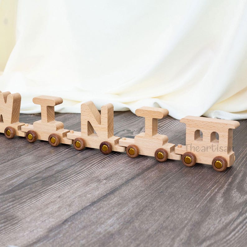 May include: A wooden toy train spelling out the name "MINI". The train is made of light-colored wood with gold-colored wheels. The train is on a wooden surface.
