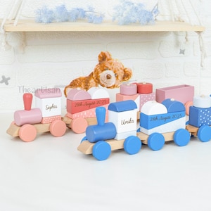 May include: A pastel-coloured wooden toy train set, featuring pink, blue, and white carriages, each with personalised names and dates. The train cars are linked and sit on a wooden track. A teddy bear is in the background.