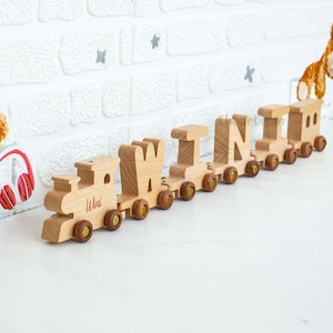 May include: A wooden toy train with the name "Wini" spelled out in individual letter cars. The train is made of light-colored wood with brown wheels and accents. The train is on a white surface with a white brick wall in the background.