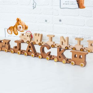 May include: Wooden train set spelling out the name "Jack" in a mix of light and dark wood. Each letter is a separate train car, connected by small yellow wheels. The background features a white brick wall.