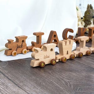May include: Wooden toy train set with individual cars spelling out names. The train cars are light and dark brown, with gold-colored wheels. The names "Jack" and "Wini" are engraved on the train cars.