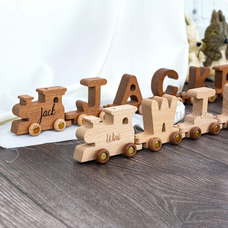 May include: Wooden toy train set with individual cars spelling out names. The train cars are light and dark brown, with gold-colored wheels. The names "Jack" and "Wini" are engraved on the train cars.