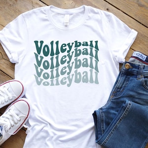 Könnte beinhalten: Weißes T-Shirt mit dem Wort "Volleyball" in einem sich wiederholenden, wellenförmigen Design in Türkistönen. Das Shirt wird mit blauen Jeans und weißen Turnschuhen auf einer Holzoberfläche präsentiert.