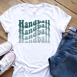 Könnte beinhalten: Weißes T-Shirt mit dem Wort "Handball" in einem wellenförmigen, mehrschichtigen Design in Türkis- und Blautönen. Das Shirt wird mit blauen Jeans und weißen Turnschuhen kombiniert, was einen lässigen, sportlichen Stil ergibt.