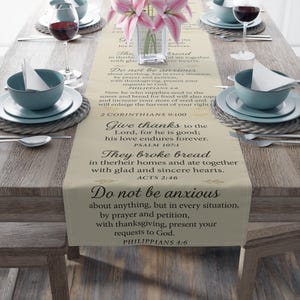 May include: A beige table runner featuring black printed Bible verses including Philippians 4:6, 2 Corinthians 9:10, Psalm 107:1, and Acts 2:46. The runner is centered on a wooden table with place settings consisting of blue bowls, plates, and wine glasses.