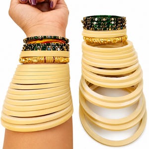 Op de afbeelding: Een verzameling gouden en crèmekleurige armbanden. De afbeelding toont een pols versierd met meerdere armbanden, waaronder gouden en groen gebeitelde ontwerpen. De armbanden zijn gestapeld, waardoor een gelaagd effect ontstaat. De achtergrond is effen wit.