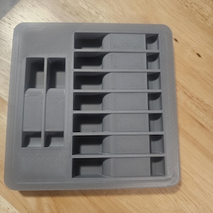 May include: A gray, rectangular organizer with multiple compartments. The organizer has a series of rectangular slots on one side and several smaller, square compartments on the other. The item is made of a solid, matte material.