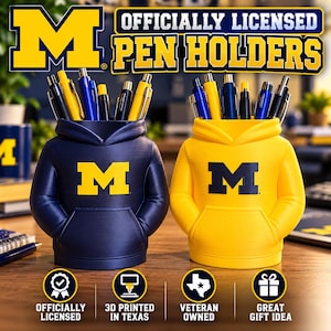 May include: Two hoodie-shaped pen holders featuring the University of Michigan block M logo. One holder is navy blue with a yellow logo, and the other is yellow with a navy blue logo. Both are filled with various pens and pencils.