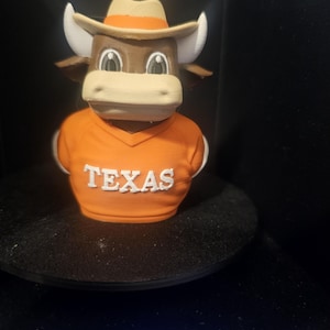 May include: A 3D-printed figurine of a Longhorn steer wearing an orange shirt with "TEXAS" in white block letters. The steer has a brown face, white horns, and a tan cowboy hat. The figurine is on a black surface.
