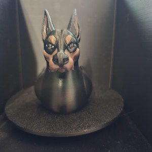 May include: A 3D-printed Doberman Pinscher figurine. The dog has a black body with brown and tan markings on its face and chest. The figurine is sitting on a dark gray circular base, with a black background.