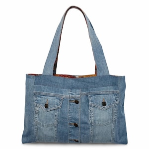 Puede incluir: Un bolso tote de mezclilla hecho de una chaqueta vaquera reutilizada. El bolso presenta la parte delantera de la chaqueta con bolsillos y cierres de botones. El forro interior tiene un diseño estampado. El bolso tiene dos correas de mezclilla para llevar.
