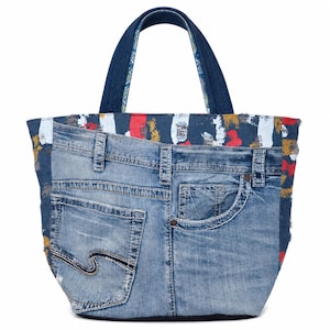 Puede incluir: Un bolso tote de mezclilla hecho de jeans reciclados. El bolso presenta los bolsillos traseros y las costuras de los jeans, con un asa de mezclilla azul. El borde superior tiene un diseño pintado colorido en rojo, blanco y amarillo.