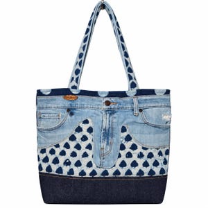 Puede incluir: Un bolso tote confeccionado con tela de mezclilla azul claro y oscuro. El diseño incorpora características de jeans, incluyendo bolsillos, bragueta y pretina. El bolso presenta un panel de tela con estampado blanco y azul y dos correas a juego.