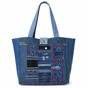Puede incluir: Un bolso tote de mezclilla azul con diseño de parches, bordados florales en rojo y azul cerceta, tiras de mezclilla cosidas horizontalmente, un panel negro texturizado y un cierre central a presión. Las letras PR están bordadas en la esquina superior derecha.