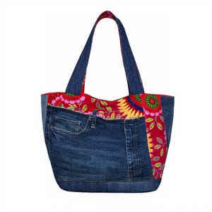 Puede incluir: Un bolso tote de mezclilla con una parte superior y detalles estampados florales rojos. La bolsa presenta un bolsillo de mezclilla y asas de mezclilla. El estampado floral incluye colores rosa, amarillo y verde.