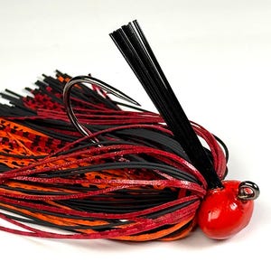 May include: A fishing jig with a red, teardrop-shaped head and a large, curved hook. The jig features a skirt of black, red, and orange rubber strands. A cluster of black, rigid strands extends from the top.