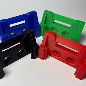 May include: Four 3D-printed brackets in blue, green, red, and black. Each bracket has a rectangular opening and two circular holes. The brackets are designed to hold or support another object.