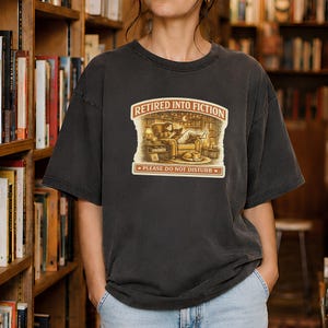 Op de afbeelding: Een antracietgrijs t-shirt met een grafische print van een persoon die in een fauteuil leest met een kat in de buurt. De tekst "RETIRED INTO FICTION" staat bovenaan en "PLEASE DO NOT DISTURB" onderaan, in een rechthoekige omlijsting.