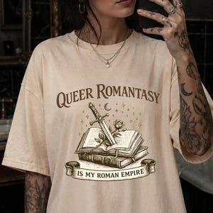 May include: A beige short-sleeve t-shirt featuring a dark brown graphic print. The design displays an open book with a sword and a rose resting on it, surrounded by stars and a crescent moon. The text reads "QUEER ROMANTASY IS MY ROMAN EMPIRE."