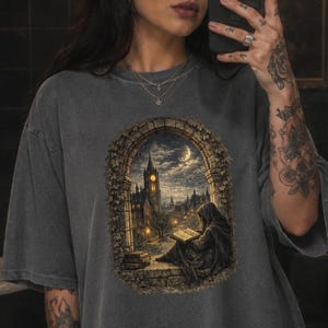 Könnte beinhalten: Ein anthrazitfarbenes Kurzarm-T-Shirt mit einem Grafikdruck einer Kapuzenfigur, die auf einem Steinvorsprung ein Buch liest. Die Szene wird von einem Steinbogen eingerahmt, der den Blick auf eine gotische Stadt bei Nacht freigibt.