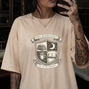 May include: A beige t-shirt featuring a black ink-style graphic of a shield divided into four quadrants. The shield displays a stack of books, a crescent moon, a steaming mug, and an open book. Text reads FERAL READERS SOCIETY and EST. IN THE MARGINS.