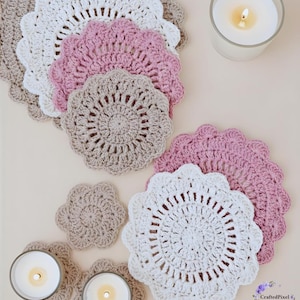 May include: A collection of handmade crochet coasters in various colors, including white, pink, and beige. The coasters have a scalloped edge design and are arranged with lit candles in glass holders. The image is taken from an overhead perspective.