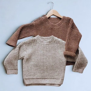 Op de afbeelding: Twee gebreide truien in de afbeelding. De bovenste trui is warmbruin en hangt aan een houten hanger. De onderste trui is lichtbeige. Beide truien hebben een gestructureerd, wafelachtig breipatroon en lange mouwen.