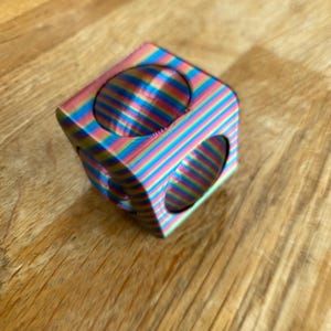 May include: A multi-coloured cube with rounded edges and circular cutouts. The cube has a striped pattern in shades of blue, pink, yellow, and green. The cube is on a wooden surface.